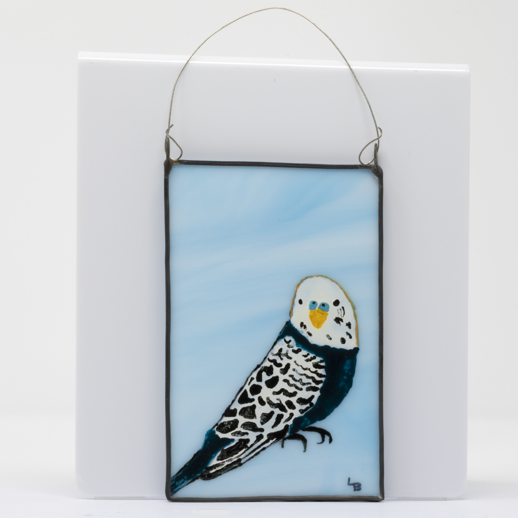 A small wall plaque comprising a hand-painted budgerigar on a background of pale blue stained glass, mounted in a dark grey lead frame with a wire hanger.