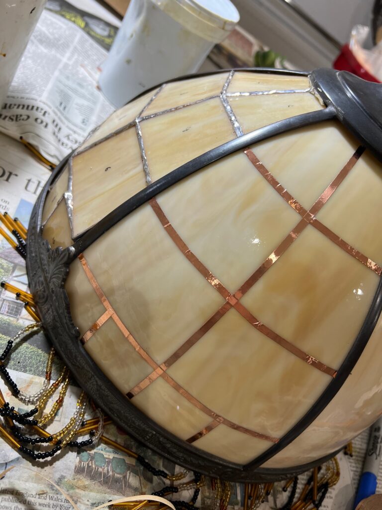 Close-up of Tiffany-style lamp repair showing original curved glass panels mounted in a heavy brown metal frame. Next to the undamaged curved panel is a glass panel made from 10 pieces of flat glass soldered together in a grid formation to create a new curve. On the undamaged curved glass copper foil strips have been applied to marry up with the lines of the replaced flat pieces. Work in progress.
