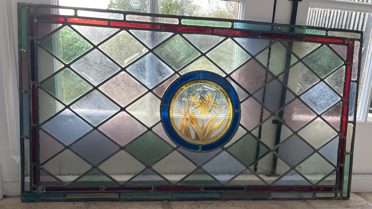 A horizontal orientation rectangular Victorian stained glass window standing against patio doors with light coming from behind. Featuring a narrow red border and horizontal diamonds with a centre painted iris in a circular blue surround.